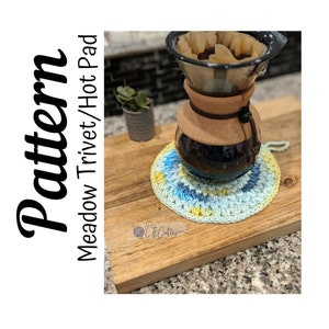 May include: A crocheted hot pad in shades of blue, yellow, and green, with a coffee maker on top. The hot pad is circular and sits on a wooden cutting board. The image includes the text "Pattern Meadow Trivet/Hot Pad".