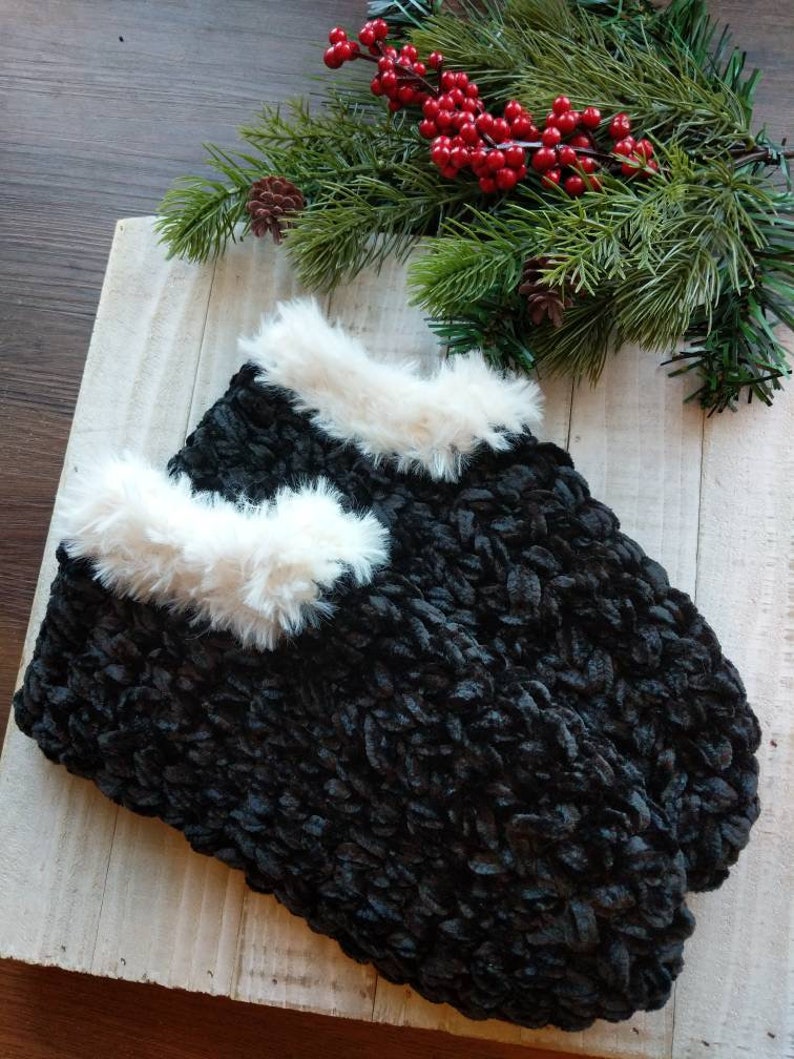 May include: A pair of black, knitted slippers with a fluffy, white trim. The slippers are displayed on a light-colored wooden surface, with evergreen branches and red berries in the background, suggesting a winter theme.
