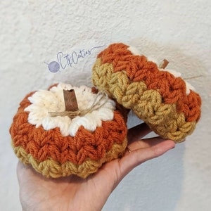 May include: Two handmade crochet pumpkins in shades of orange, cream, and yellow. Each pumpkin has a cinnamon stick stem and twine detail. The pumpkins are a fall-themed decoration.