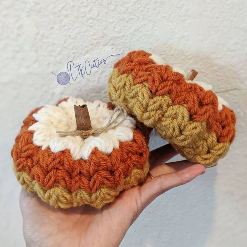 May include: Two handmade crochet pumpkins in shades of orange, cream, and yellow. Each pumpkin has a cinnamon stick stem and twine detail. The pumpkins are a fall-themed decoration.
