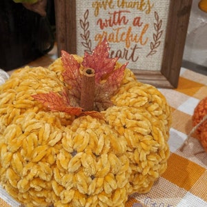 May include: A plush, yellow pumpkin with a cinnamon stick stem and decorative leaves. A framed sign in the background reads "give thanks with a grateful heart". The pumpkin is on a checkered orange and white surface.