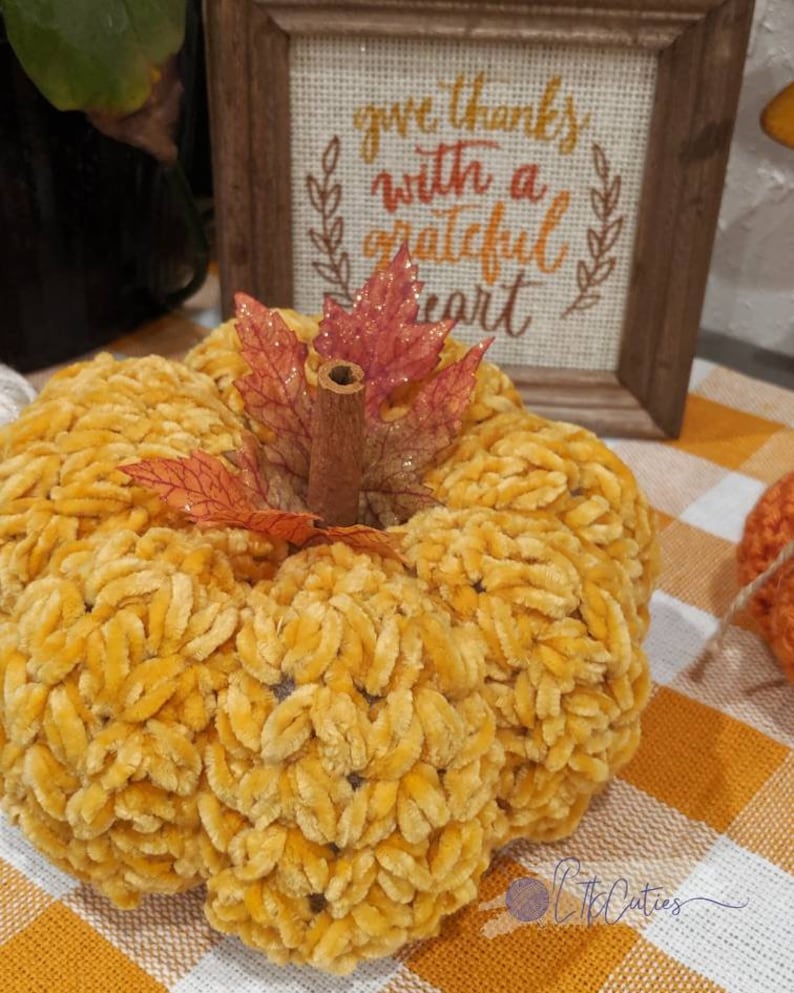 May include: A plush, yellow pumpkin with a cinnamon stick stem and decorative leaves. A framed sign in the background reads "give thanks with a grateful heart". The pumpkin is on a checkered orange and white surface.