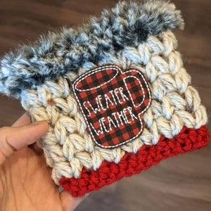 K&ouml;nnte beinhalten: Ein geh&auml;keltes Kaffeetassen-Cozy mit einem roten und schwarzen karierten Becherdesign, auf dem "Sweater Weather" steht. Das Cozy hat einen wei&szlig;en geh&auml;kelten K&ouml;rper mit einem roten geh&auml;kelten Boden und einem schwarzen und wei&szlig;en Kunstfellbesatz.