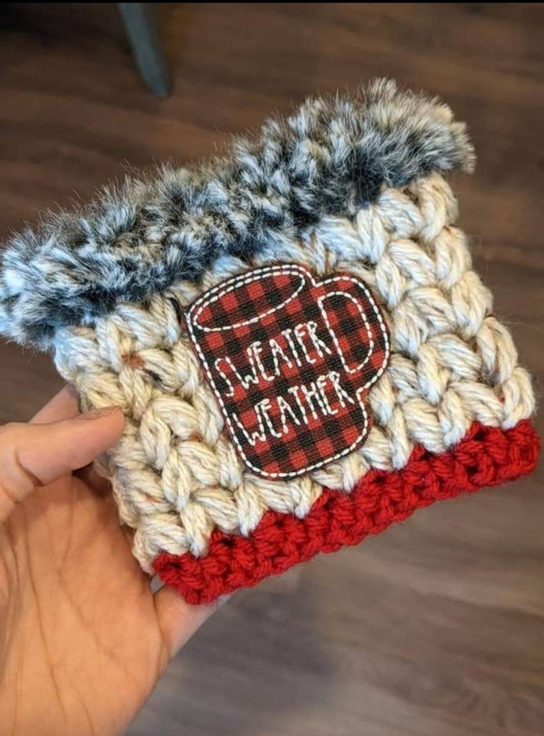 K&ouml;nnte beinhalten: Ein geh&auml;keltes Kaffeetassen-Cozy mit einem roten und schwarzen karierten Becherdesign, auf dem "Sweater Weather" steht. Das Cozy hat einen wei&szlig;en geh&auml;kelten K&ouml;rper mit einem roten geh&auml;kelten Boden und einem schwarzen und wei&szlig;en Kunstfellbesatz.