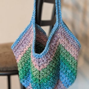 May include: A crocheted tote bag with a blue, green, and purple striped design. The bag has a wide, open top and two long straps.