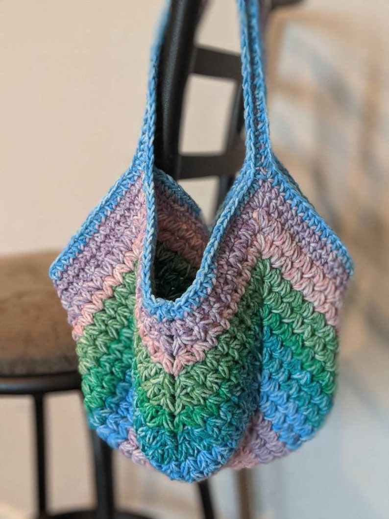 May include: A crocheted tote bag with a blue, green, and purple striped design. The bag has a wide, open top and two long straps.