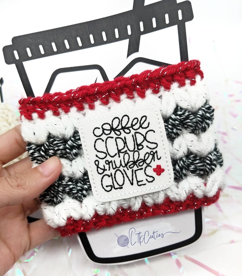K&ouml;nnte beinhalten: Rote, wei&szlig;e und schwarze geh&auml;kelte Kaffeetassen-H&uuml;lle mit einem wei&szlig;en Patch, auf dem "coffee scrubs & rubber gloves" mit einem roten Kreuz steht.