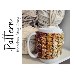 May include: A crocheted mug cozy in shades of brown, orange, and green. The cozy is on a white mug and surrounded by balls of yarn.