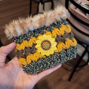 K&ouml;nnte beinhalten: Ein geh&auml;kelter Becherw&auml;rmer mit Sonnenblumenmuster. Der W&auml;rmer ist aus gr&uuml;nem, braunem und gelbem Garn gefertigt und hat einen Kunstfellbesatz.