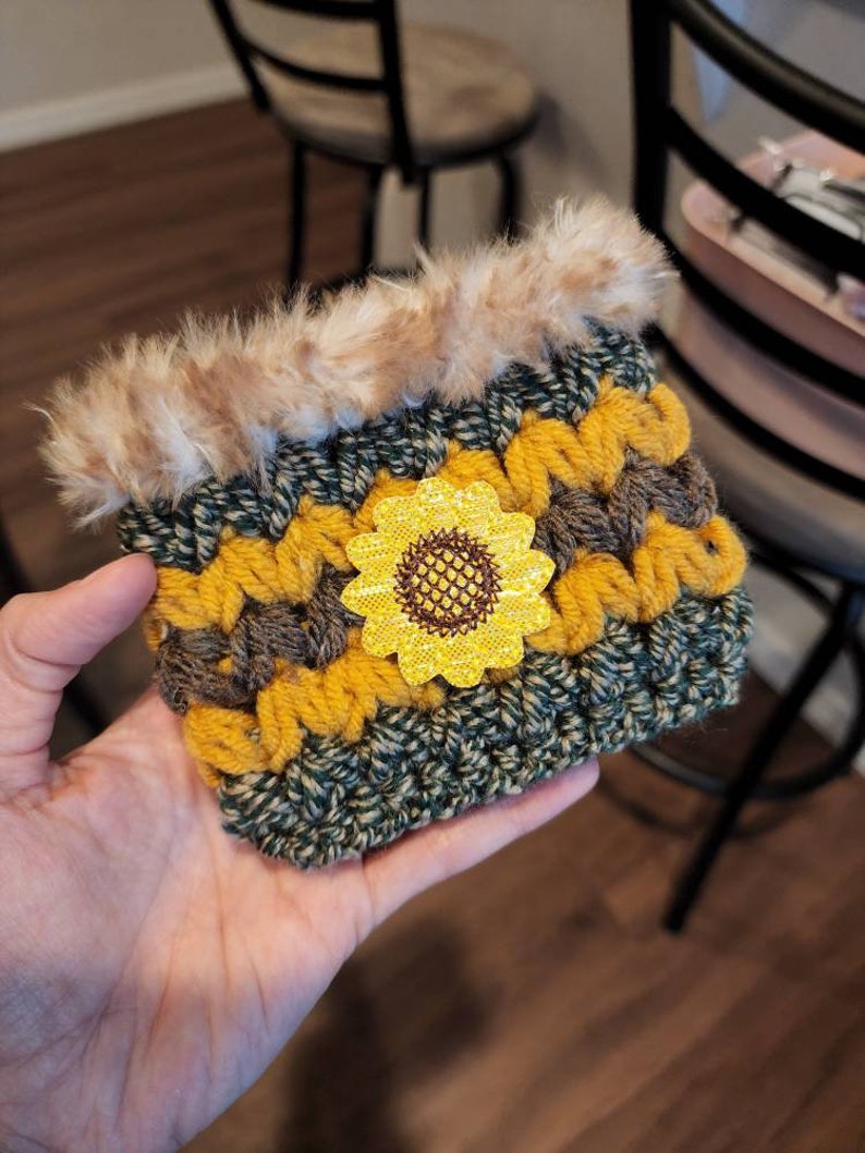 K&ouml;nnte beinhalten: Ein geh&auml;kelter Becherw&auml;rmer mit Sonnenblumenmuster. Der W&auml;rmer ist aus gr&uuml;nem, braunem und gelbem Garn gefertigt und hat einen Kunstfellbesatz.