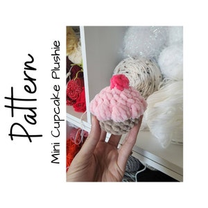 May include: A pink and brown yarn cupcake plush toy with a red cherry on top. The plush is being held by a hand in front of a shelf with balls of yarn.