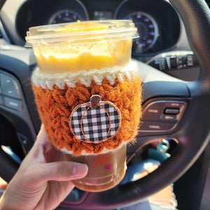 K&ouml;nnte beinhalten: Nahaufnahme eines K&uuml;rbisgew&uuml;rz-Lattes in einem durchsichtigen Becher mit einer geh&auml;kelten orangefarbenen und wei&szlig;en H&uuml;lle mit einem schwarzen und wei&szlig;en karierten K&uuml;rbisdesign.