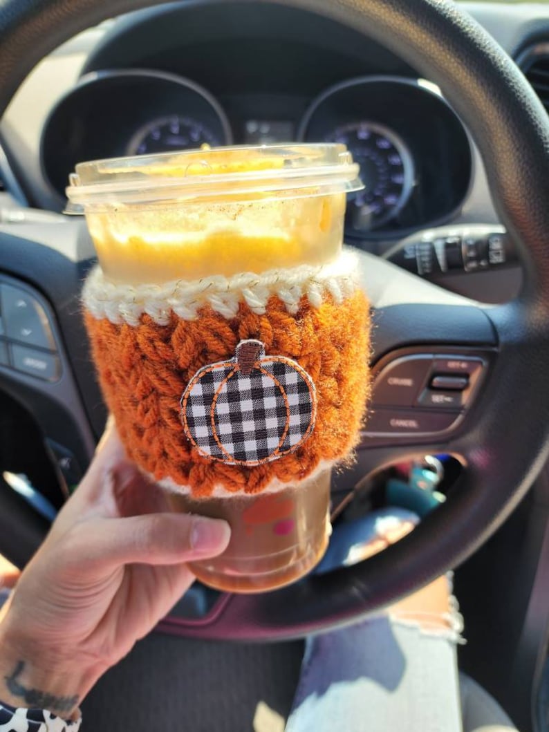 K&ouml;nnte beinhalten: Nahaufnahme eines K&uuml;rbisgew&uuml;rz-Lattes in einem durchsichtigen Becher mit einer geh&auml;kelten orangefarbenen und wei&szlig;en H&uuml;lle mit einem schwarzen und wei&szlig;en karierten K&uuml;rbisdesign.
