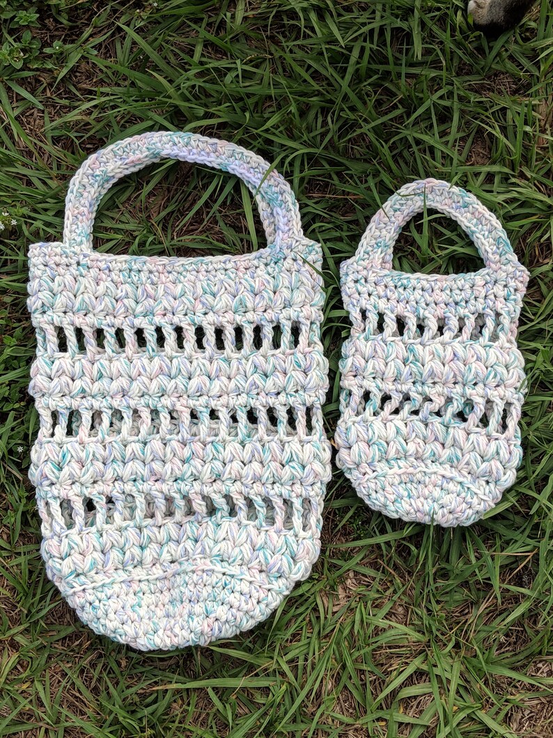 May include: Two crocheted bags made with a white yarn with blue, pink, and purple flecks. The larger bag has a wider opening and a longer handle. The smaller bag has a smaller opening and a shorter handle.
