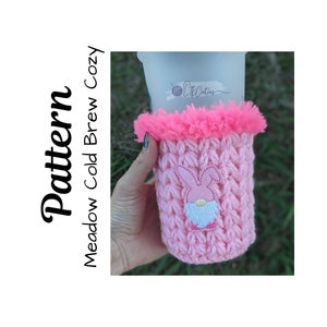 Pode incluir: Capa de crochet rosa para caneca com uma borda de pelo rosa e um design de gnomo coelho bordado. A capa diz "Little Cuties" na caneca.