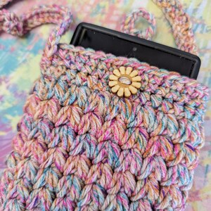May include: A crocheted phone pouch with a colorful, multi-colored yarn. The pouch has a wooden button with a flower design. The pouch is being held by a matching crocheted strap.