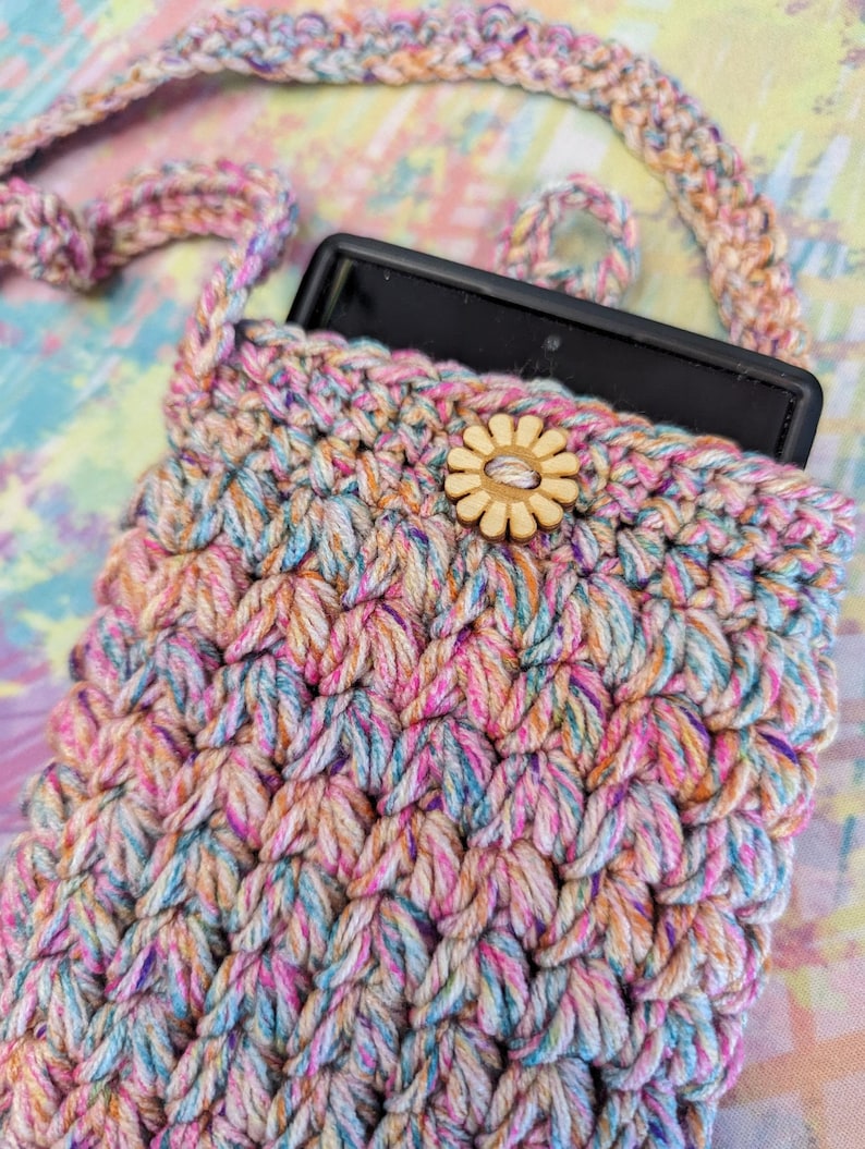 May include: A crocheted phone pouch with a colorful, multi-colored yarn. The pouch has a wooden button with a flower design. The pouch is being held by a matching crocheted strap.