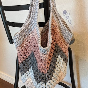 May include: A crocheted tote bag with a chevron pattern in shades of gray, pink, and white. The bag has a wide, flat bottom and two long straps.