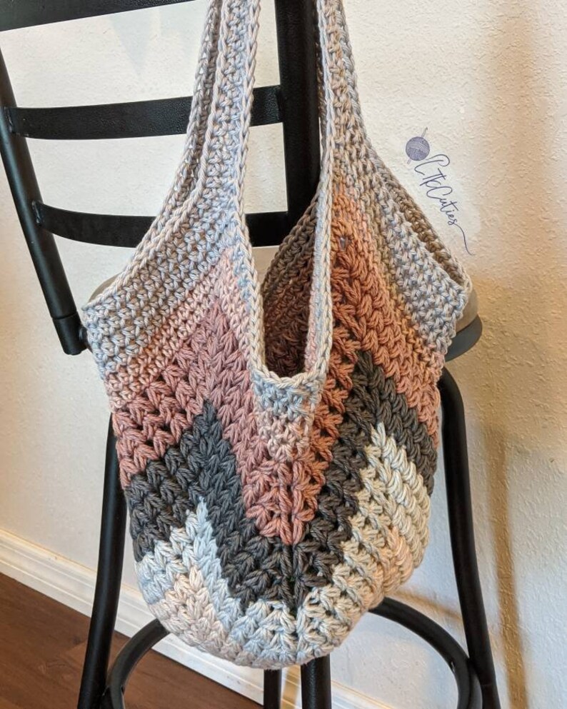 May include: A crocheted tote bag with a chevron pattern in shades of gray, pink, and white. The bag has a wide, flat bottom and two long straps.
