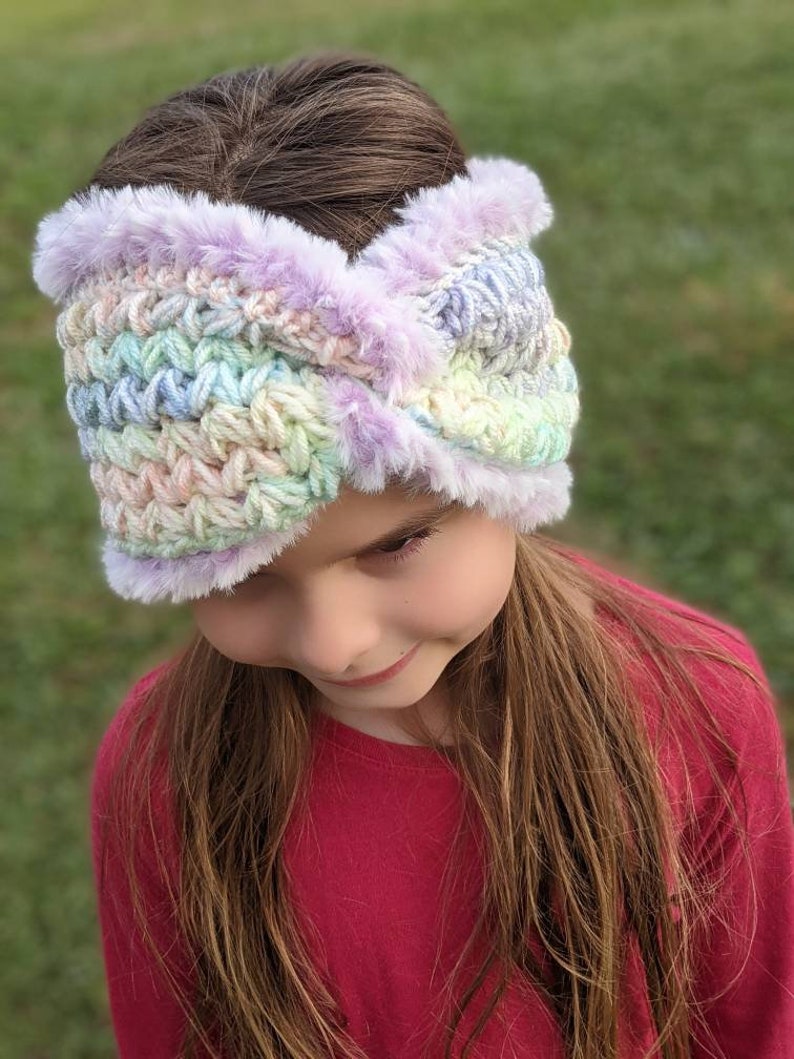 May include: A pastel-colored crochet headband with a purple faux fur trim. The headband is worn by a young person with long brown hair.