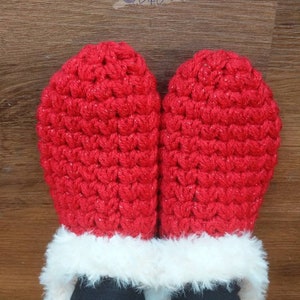 May include: A pair of red crocheted mittens with a white faux fur trim. The mittens are made of a textured yarn and have a sparkly appearance. The mittens are shown on a person wearing black pants.