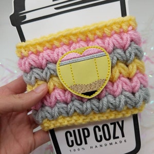 K&ouml;nnte beinhalten: Ein gelber, rosa und grauer geh&auml;kelter Becherw&auml;rmer mit einem gestickten Bleistift-Design. Der Becherw&auml;rmer ist mit "CUP COZY 100% HANDMADE" beschriftet.