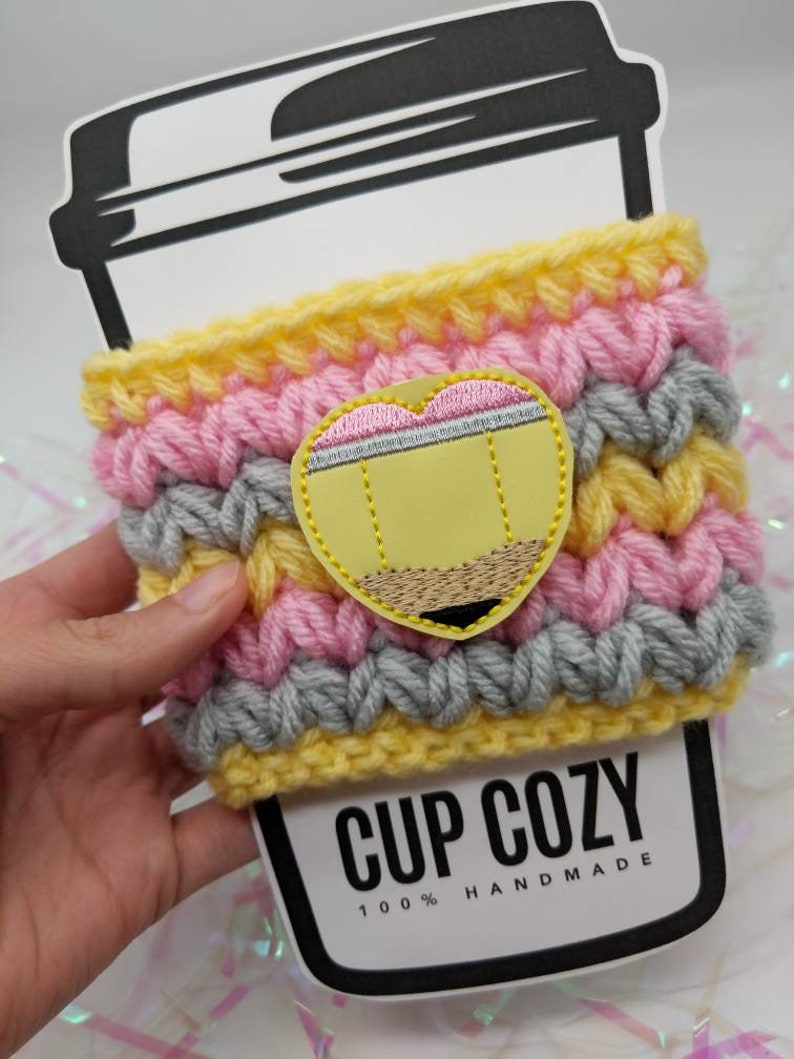 K&ouml;nnte beinhalten: Ein gelber, rosa und grauer geh&auml;kelter Becherw&auml;rmer mit einem gestickten Bleistift-Design. Der Becherw&auml;rmer ist mit "CUP COZY 100% HANDMADE" beschriftet.