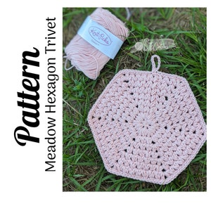 May include: A pink crochet hexagon trivet with a loop for hanging. The trivet is made with a textured stitch pattern and is lying on a bed of green grass. A ball of pink yarn is in the background.