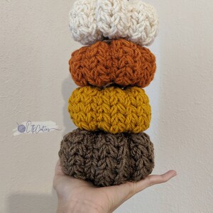 May include: A vertical stack of four crocheted pumpkins in autumnal shades: cream, orange, yellow, and brown. Each pumpkin features a textured, ribbed pattern. A twine loop is attached to the top pumpkin. These handmade pumpkins are a decorative fall accent.