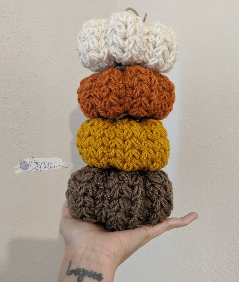 May include: A vertical stack of four crocheted pumpkins in autumnal shades: cream, orange, yellow, and brown. Each pumpkin features a textured, ribbed pattern. A twine loop is attached to the top pumpkin. These handmade pumpkins are a decorative fall accent.