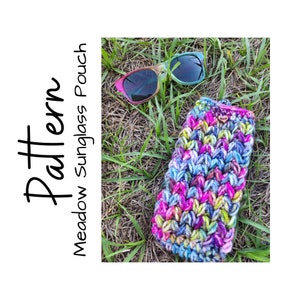 May include: A colorful crocheted pouch for sunglasses, with a bright pink, blue, and green color scheme. The pouch is lying on a bed of green grass, with a pair of sunglasses lying next to it. The text "Pattern Meadow Sunglass Pouch" is written in black on a white background.