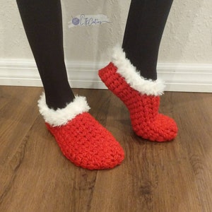 May include: Red crocheted slippers with white faux fur trim. The slippers are a festive red color and appear to be handmade. The slippers are shown on a person's feet, with black leggings and a wooden floor in the background.