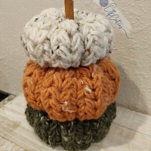 May include: A decorative stack of three crocheted pumpkins in autumnal hues. The top pumpkin is cream, the middle is orange, and the base is green. A cinnamon stick forms the stem. The pumpkins are handmade, with a textured, chunky appearance.