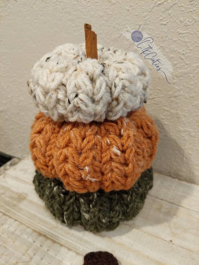 May include: A decorative stack of three crocheted pumpkins in autumnal hues. The top pumpkin is cream, the middle is orange, and the base is green. A cinnamon stick forms the stem. The pumpkins are handmade, with a textured, chunky appearance.