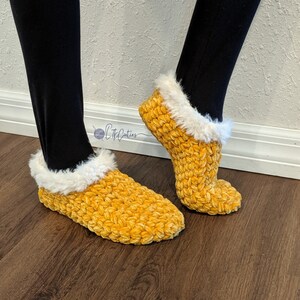 May include: A pair of yellow crochet slippers with a fluffy white trim. The slippers are made of a soft, textured yarn and are designed to be worn indoors. The slippers are shown on a person wearing black leggings.