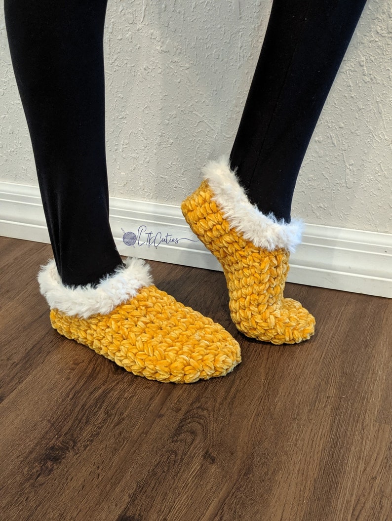 May include: A pair of yellow crochet slippers with a fluffy white trim. The slippers are made of a soft, textured yarn and are designed to be worn indoors. The slippers are shown on a person wearing black leggings.