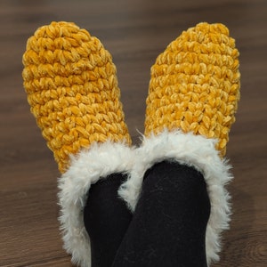 May include: A pair of cozy, mustard-yellow crochet slippers with a fluffy white trim. The slippers are made of a soft, textured yarn and are worn over black leggings. The image is taken from a low angle, showing the feet and lower legs.