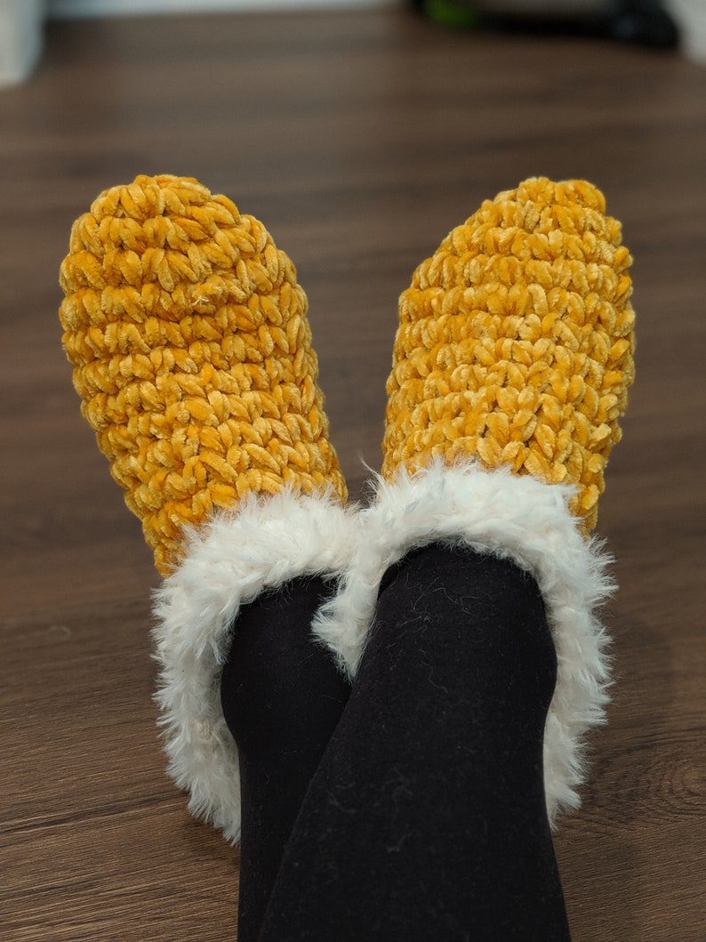 May include: A pair of cozy, mustard-yellow crochet slippers with a fluffy white trim. The slippers are made of a soft, textured yarn and are worn over black leggings. The image is taken from a low angle, showing the feet and lower legs.