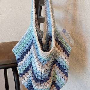 May include: A crocheted tote bag with a blue, green, and white color scheme. The bag has a wide, flat bottom and two long straps.