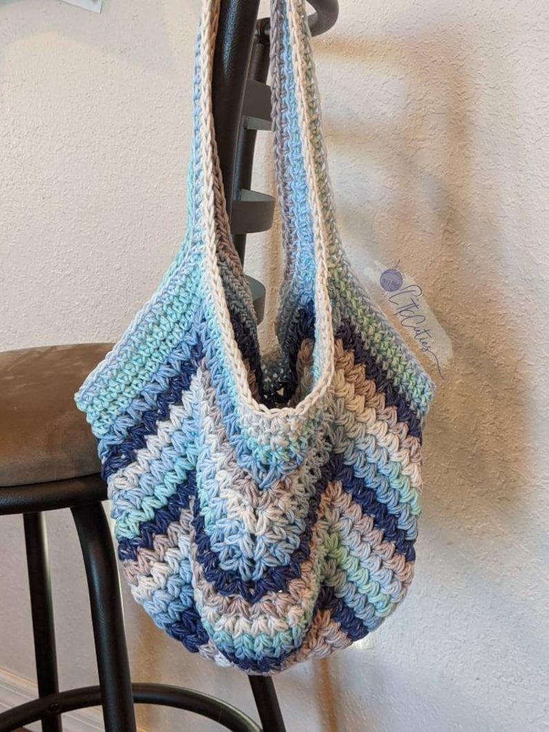 May include: A crocheted tote bag with a blue, green, and white color scheme. The bag has a wide, flat bottom and two long straps.