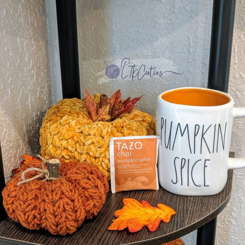 May include: A seasonal display with two crocheted pumpkins in orange and yellow, a white mug with "PUMPKIN SPICE" text, and a Tazo Chai Pumpkin Spice tea packet. Artificial autumn leaves add to the autumnal theme.