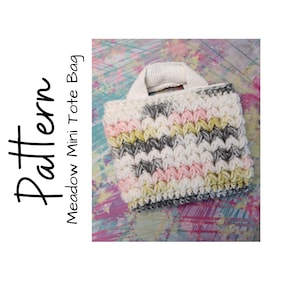 May include: A small, hand-crocheted tote bag with a textured stitch pattern. The bag features a color palette of white, pink, yellow, and gray stripes. The handle is a solid white. The text "Pattern Meadow Mini Tote Bag" is on the left side.