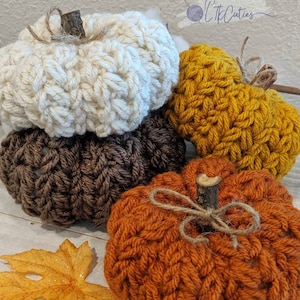 May include: Four handmade crochet pumpkins in autumnal shades: cream, brown, mustard, and orange. Each pumpkin has a twig stem and twine bow. An orange leaf is in the foreground. These pumpkins are a cosy autumn decoration.