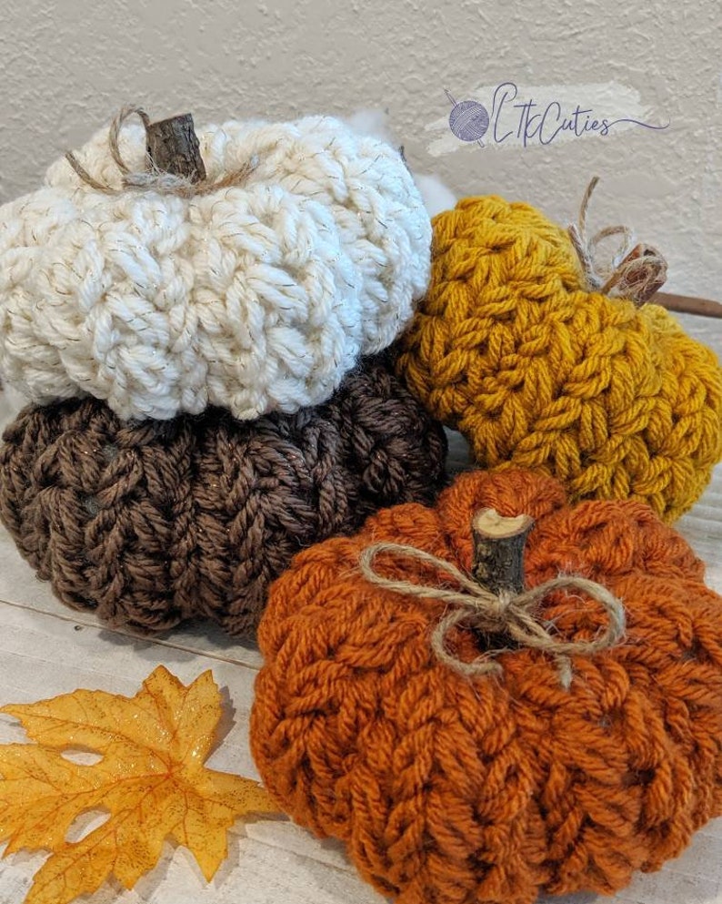 May include: Four handmade crochet pumpkins in autumnal shades: cream, brown, mustard, and orange. Each pumpkin has a twig stem and twine bow. An orange leaf is in the foreground. These pumpkins are a cosy autumn decoration.