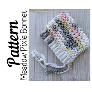 May include: A crocheted baby bonnet in shades of pink, white, and gray with a matching crocheted tie. The bonnet is called "Meadow Pixie Bonnet" and is shown on a wooden surface.