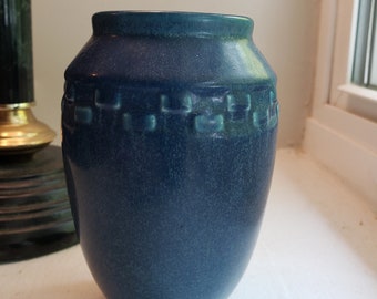 May include: A blue ceramic vase with a textured surface and a band of square designs around the top. The vase has a slightly flared rim and a rounded body. The color is a deep blue with hints of teal.