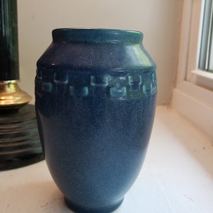 May include: A blue ceramic vase with a textured surface and a band of square designs around the top. The vase has a slightly flared rim and a rounded body. The color is a deep blue with hints of teal.