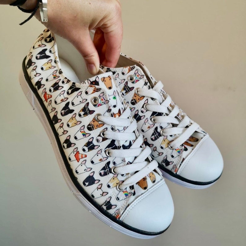 May include: White canvas low-top sneakers with a black trim and white laces. The sneakers are covered in a pattern of colorful bull terrier dog faces.