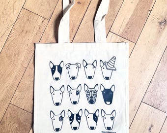 Puede incluir: Un bolso de tela color crema con un estampado gráfico negro de doce caras de perro Bull Terrier en un patrón de cuadrícula. Un perro lleva un cono de tráfico en la cabeza. El texto emilydevineart está impreso en letras negras en la parte inferior.