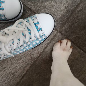 May include: A pair of white canvas shoes with a blue and white pattern of small dogs. The shoes are tied with white laces. A white dog's paw is visible in the lower right corner of the image.